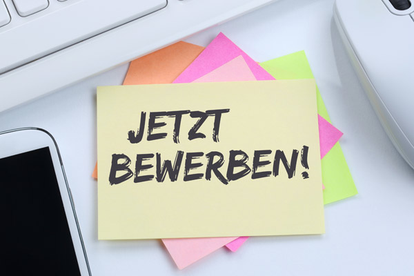 Ein gelbes Haftnotiz mit der Aufschrift JETZT BEWERBEN auf einem Stapel von bunten Haftnotizen, neben einem Smartphone und einer Computermaus.
