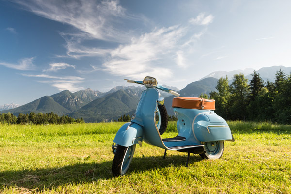 Ein blauer Roller steht auf einer Wiese mit Bergen im Hintergrund und einem klaren Himmel.